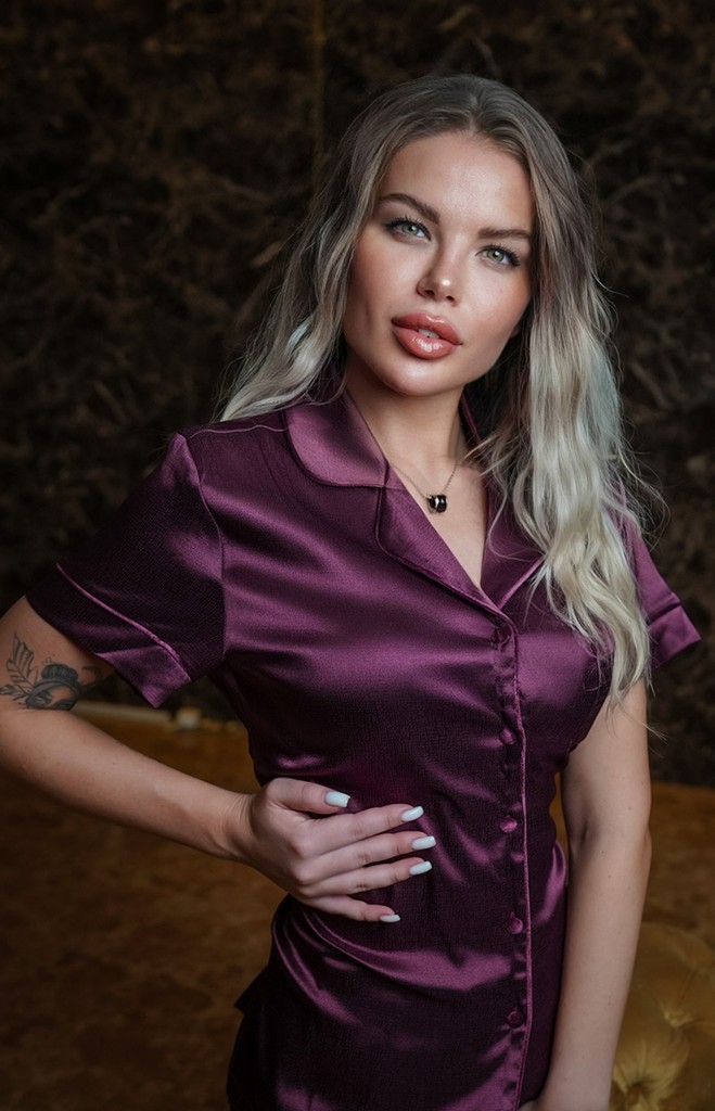 Young massage therapist with blonde hair in purple satin shirt, necklace, and arm tattoo